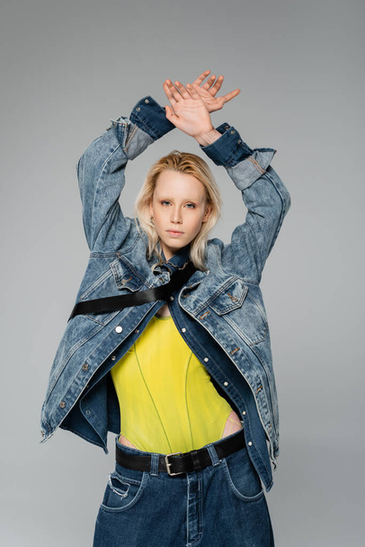 young blonde woman in stylish denim outfit looking at camera while posing with hands above head isolated on grey  - Foto, immagini