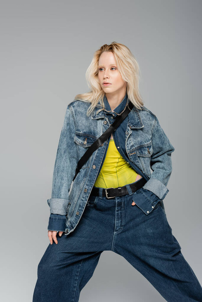 young blonde woman in stylish denim clothes posing and looking away isolated on grey  - Foto, Bild