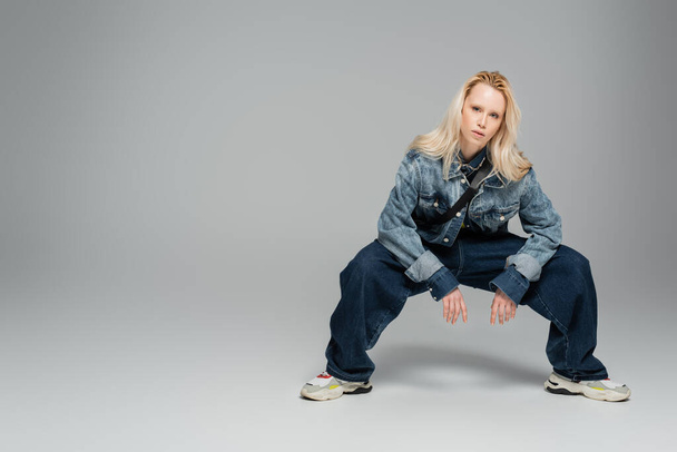 повна довжина молодої блондинки в стильному джинсовому одязі позує на сірому
  - Фото, зображення