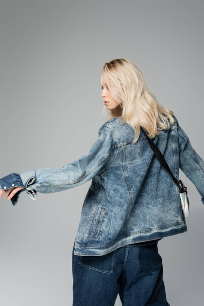 young blonde woman in stylish denim jacket with belt bag posing isolated on grey  - Φωτογραφία, εικόνα