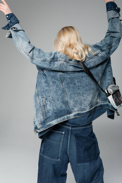 πίσω όψη του νεαρού ξανθό μοντέλο σε κομψό τζιν σακάκι με τσάντα ζώνη που θέτουν με υψωμένα χέρια απομονώνονται σε γκρι  - Φωτογραφία, εικόνα