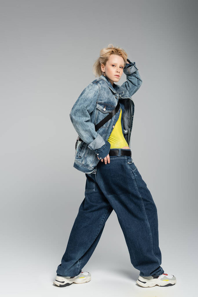 full length of young blonde model in stylish denim outfit and trendy sneakers posing on grey  - Photo, Image