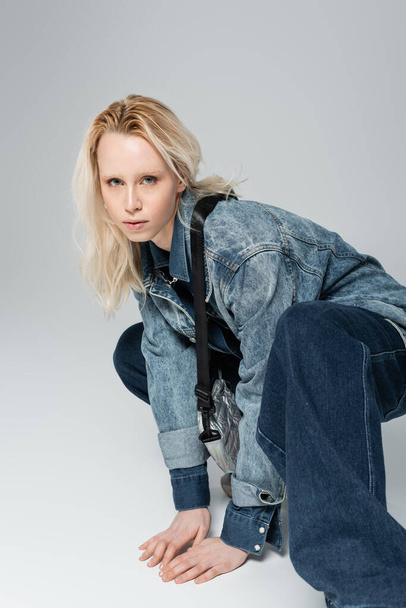 блондинка альбіно модель в стильному джинсовому вбранні з сумкою для пояса позує на сірому
  - Фото, зображення
