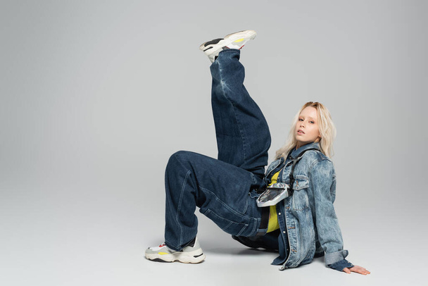 full length of young blonde model in denim outfit and trendy sneakers posing on grey  - Foto, Bild