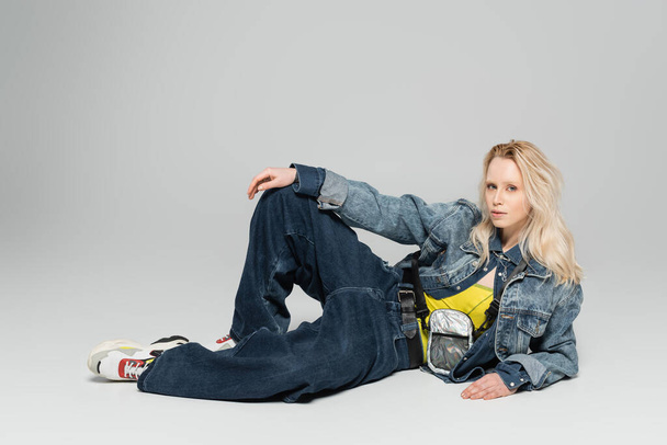 full length of young blonde woman in blue denim outfit and trendy sneakers posing on grey background  - Foto, imagen