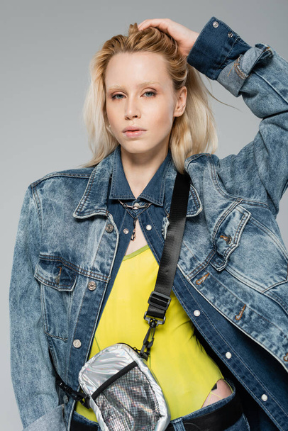 young woman in stylish denim jacket posing while adjusting blonde hair isolated on grey  - Фото, изображение
