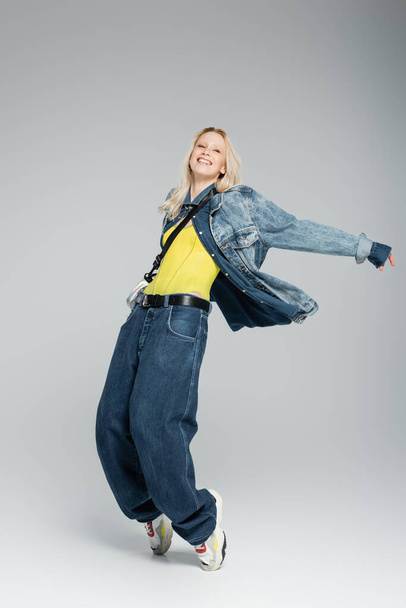 longitud completa de la joven feliz en traje de mezclilla azul y elegantes zapatillas de deporte posando en gris  - Foto, imagen