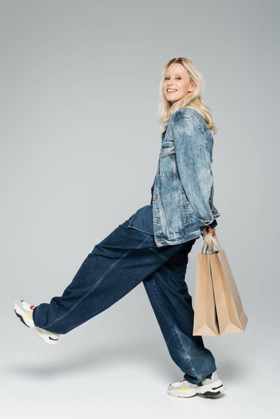 volledige lengte van vrolijke jonge vrouw in stijlvolle denim outfit met winkeltassen en wandelen op grijs  - Foto, afbeelding