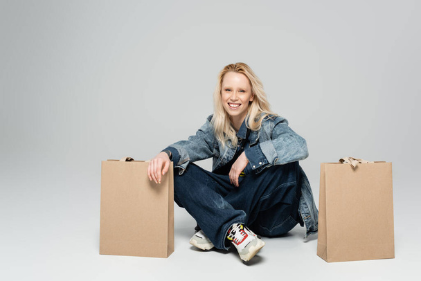 повна довжина радісної молодої жінки в стильному джинсовому вбранні, що сидить біля сумки для покупок на сірому
  - Фото, зображення