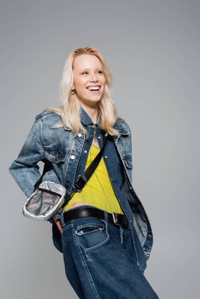 happy woman in denim outfit and belt bag smiling isolated on grey  - Foto, Imagen