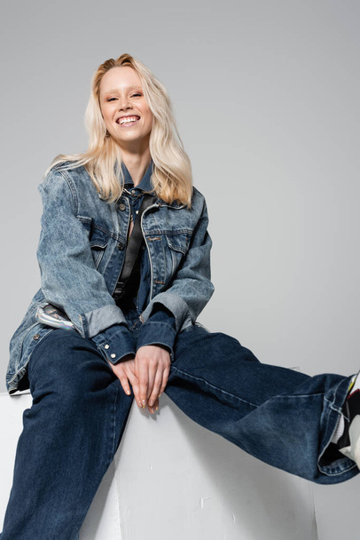 happy blonde woman in blue denim outfit and trendy jeans sitting on white cube isolated on grey  - Foto, Bild