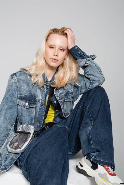 blonde woman in blue denim outfit and trendy jeans sitting on white cube isolated on grey  - Foto, Bild