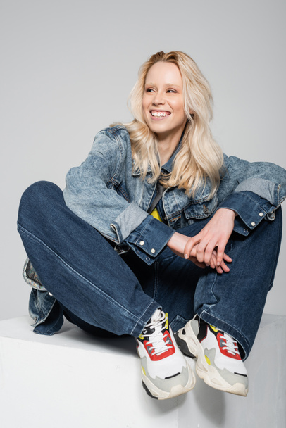 full length of happy blonde woman in blue denim outfit and trendy sneakers sitting on white cube isolated on grey  - Foto, Imagen