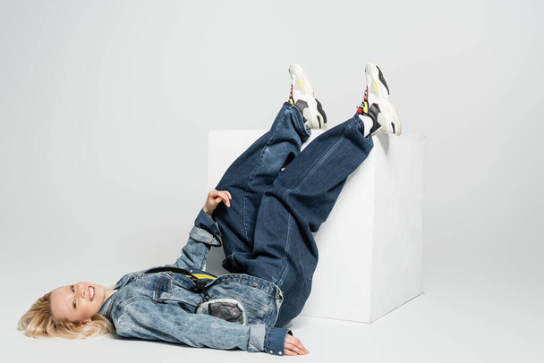 full length of cheerful blonde woman in blue denim outfit and trendy sneakers lying near white cube on grey  - Foto, afbeelding
