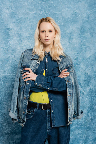 blonde model in denim jacket posing with crossed arms near blue textured background   - 写真・画像