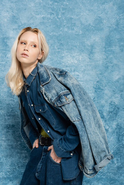 blonde woman in denim jacket posing with hands in pockets near blue textured background   - Foto, immagini