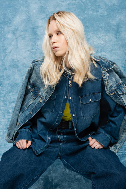blonde woman in denim jacket posing and looking away near blue textured background   - 写真・画像
