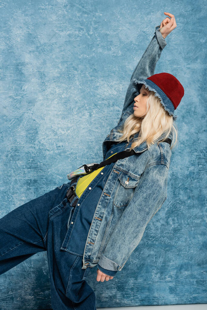 blonde woman in denim jacket and panama hat looking away and posing with raised hand near blue textured background   - Fotografie, Obrázek