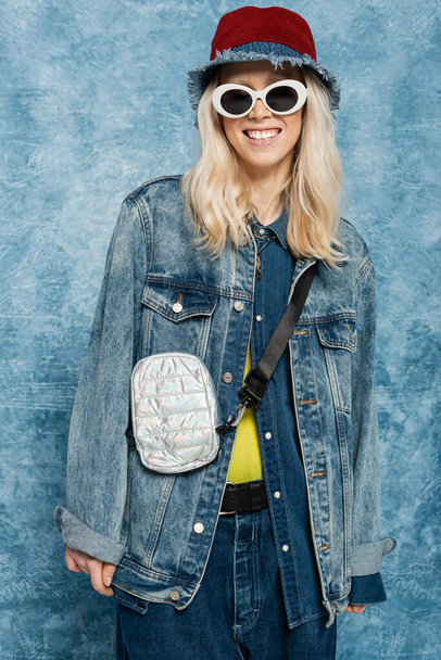 счастливая блондинка в джинсовой одежде позирует в панамской шляпе и солнечных очках рядом с синим текстурированным фоном   - Фото, изображение