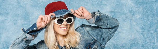 smiling blonde woman in denim jacket and sunglasses adjusting panama hat near blue textured background, banner  - Foto, afbeelding