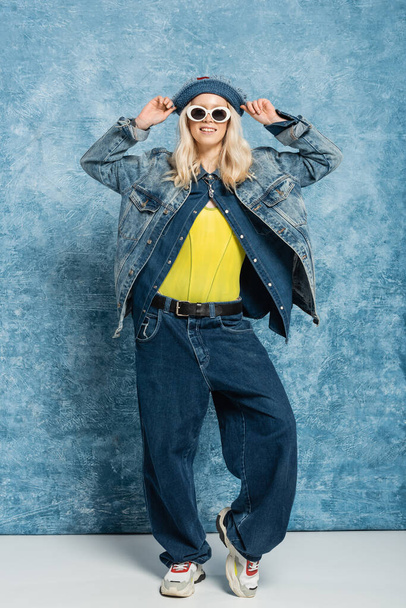 полная длина счастливая блондинка в джинсовой шляпе Панама и солнцезащитные очки, позирующие вблизи синего текстурированного фона   - Фото, изображение