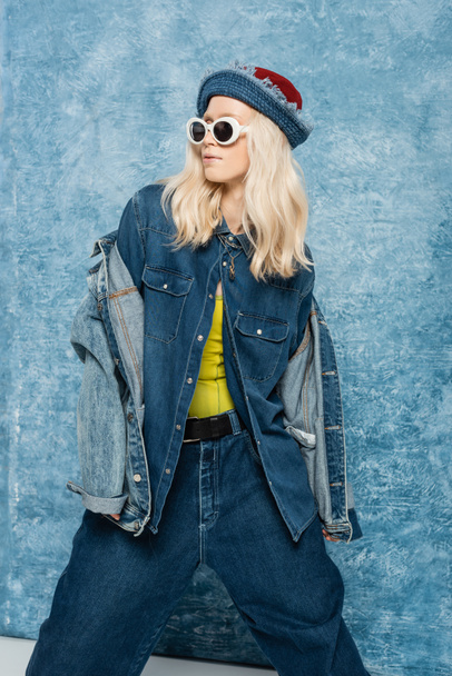 blonde woman in denim panama hat and sunglasses posing near blue textured background   - Foto, afbeelding