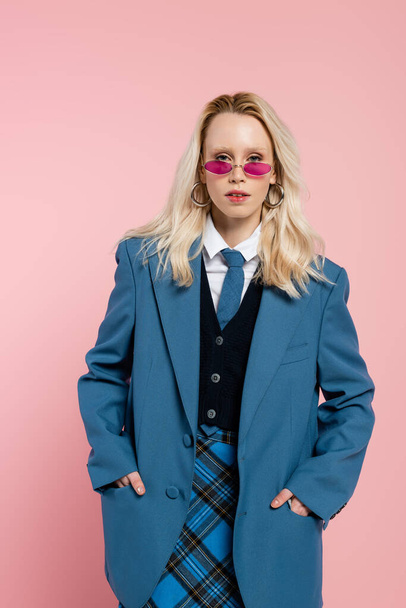 blonde woman in blue blazer with tie and sunglasses posing with hands in pockets isolated on pink  - Foto, imagen