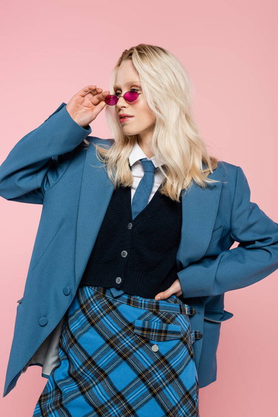 blonde woman in blue blazer with tie and sunglasses posing with hand on hip isolated on pink  - Foto, afbeelding