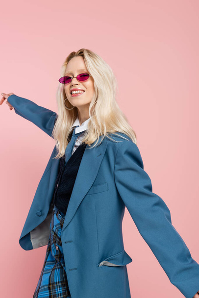 smiling woman in blue blazer with tie and sunglasses posing with outstretched hand isolated on pink  - Фото, изображение