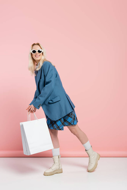 full length of happy woman in blue outfit with plaid skirt and trendy sunglasses posing with shopping bags on pink  - Фото, изображение