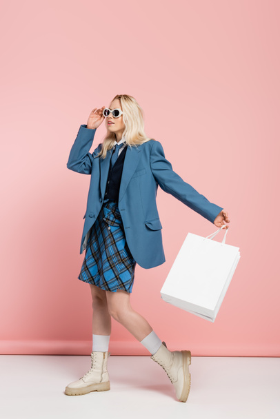 full length of blonde woman in blue blazer adjusting trendy sunglasses and holding shopping bags on pink  - Photo, Image