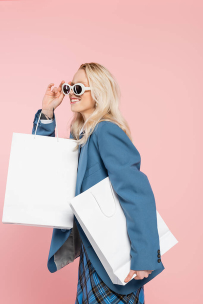 cheerful woman in blue blazer adjusting trendy sunglasses and holding shopping bags isolated on pink - Fotó, kép