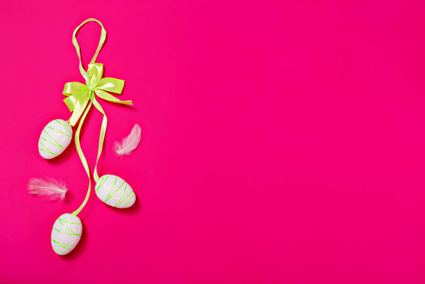 White artificial Easter eggs on light green ribbon on pink background. Nearby fluffy chicken feathers - Foto, Imagem