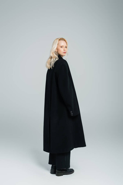 πλήρες μήκος της ξανθιάς νεαρή γυναίκα σε μαύρο παλτό στέκεται και κοιτάζοντας κάμερα σε γκρι  - Φωτογραφία, εικόνα