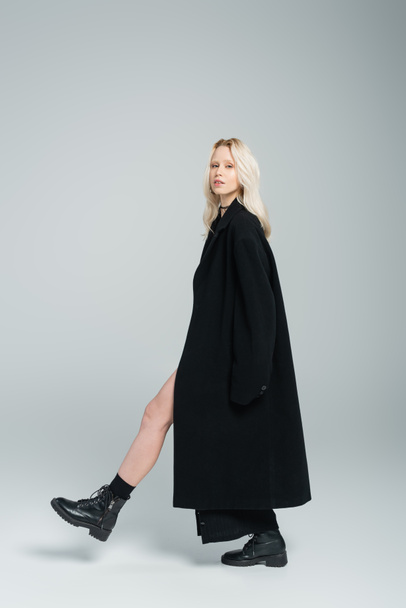 full length of blonde young woman in black coat walking and looking at camera on grey  - Foto, Imagem