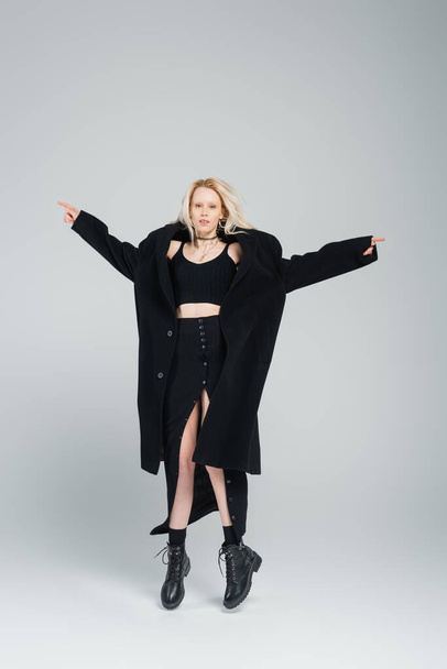 full length of blonde and young woman in stylish black outfit levitating on grey  - Fotó, kép