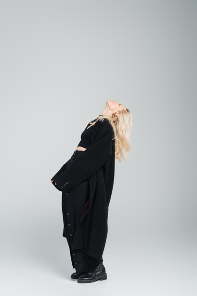 full length of blonde young woman in stylish black outfit standing with raised head on grey  - Fotó, kép