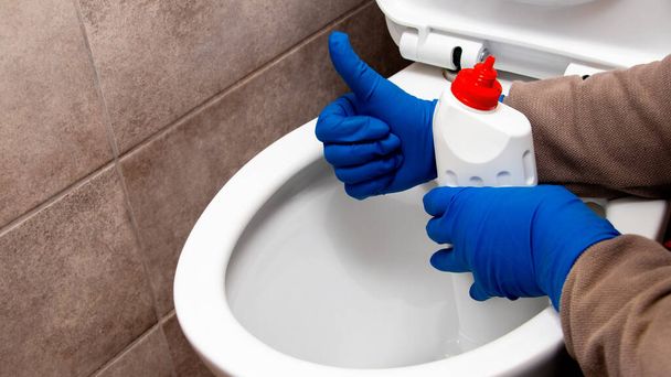 A senhora da limpeza perto da casa de banho com agente de limpeza mostra o sinal ok com a mão. A sanita está limpa. - Foto, Imagem