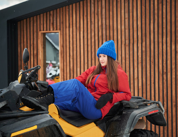 Retrato de una hermosa mujer posando en todoterreno ATV de cuatro ruedas con casa de madera en el fondo. Concepto de ocio activo y actividades invernales. - Foto, Imagen