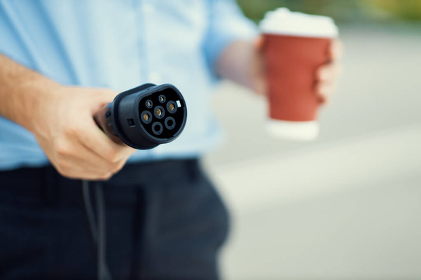Focus on EV charging plug device from charging station for electric vehicle with blurred man holding plug and pointing at the camera background for alternative renewable energy. - Photo, image