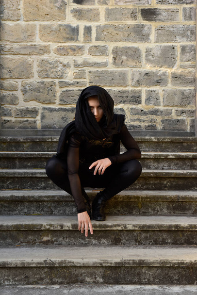 Πορτρέτο του όμορφου γυναικείου μοντέλου με ξανθιά πλεξούδα, φορώντας μαύρη δερμάτινη γατοστολή και ρέουσα κουκούλα μανδύα, φανταστικός πολεμιστής δολοφόνος. Ανάρτηση σε φόντο κάστρου με πέτρινη σκάλα. - Φωτογραφία, εικόνα