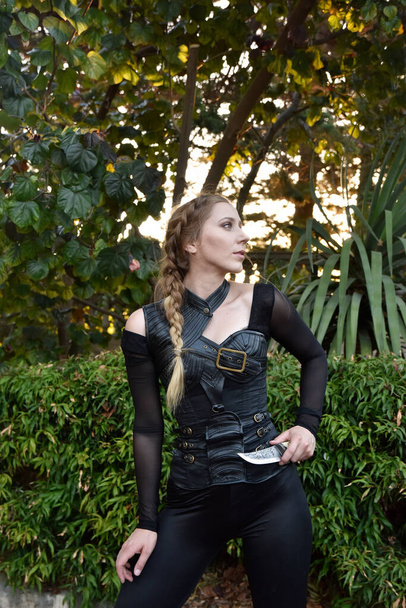 Close up portrait of beautiful female model with blonde plait, wearing black leather catsuit, fantasy assassin warrior.  Posing in forest  tree background - Photo, image