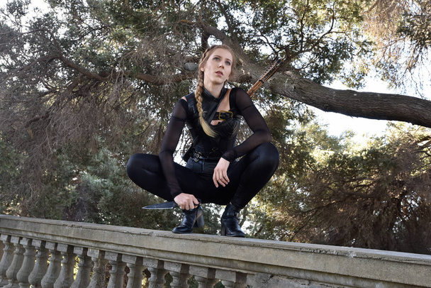Portrait of beautiful female model with blonde plait, wearing black leather catsuit costume, fantasy assassin warrior.  Crouching sitting pose, holding knife on stone  balcony of  castle background - Foto, afbeelding
