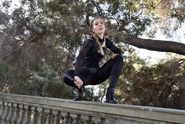 Portrait of beautiful female model with blonde plait, wearing black leather catsuit costume, fantasy assassin warrior.  Crouching sitting pose, holding knife on stone  balcony of  castle background - Foto, afbeelding