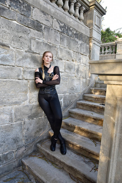Full length portrait of beautiful female model with blonde plait, wearing black leather catsuit costume, fantasy assassin warrior.  Standing walking pose on stone staircase of  castle background - 写真・画像