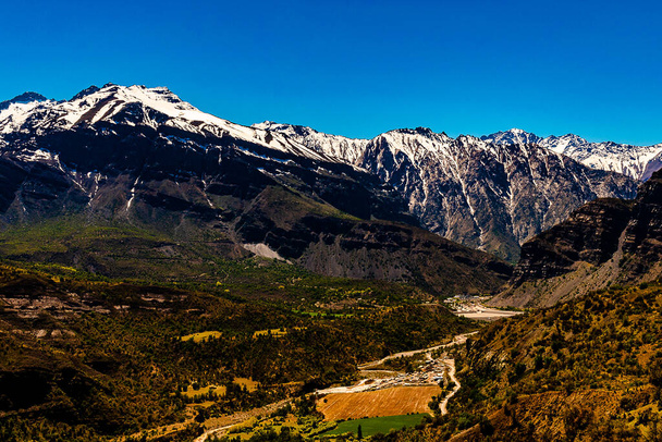 Общий вид горной долины со склерофильной растительностью, с рекой в центре и заснеженными горами на заднем плане, под ясным голубым небом - Фото, изображение
