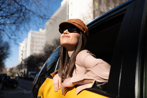 Κορίτσι γέρνει έξω από το παράθυρο ενός ταξί στη Βαρκελώνη, χαμογελαστή γυναίκα κοιτάζοντας την πόλη με ενθουσιασμό, Καυκάσιος με μακριά ίσια μαλλιά, δερμάτινο μπερέ και ανοιχτό ροζ σακάκι. - Φωτογραφία, εικόνα