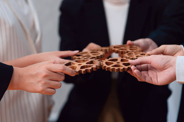 Closeup hand holding wooden gear by businesspeople wearing suit for harmony synergy in office workplace concept. Group of people hand making chain of gears into collective form for unity symbol. - Foto, afbeelding
