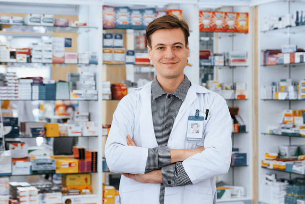 Portrait of a qualified, friendly male pharmacist wearing a white coat, crossing his arms, and looking at the camera, with a shelf of various medicine boxes in background at drugstore or pharmacy. - Photo, Image