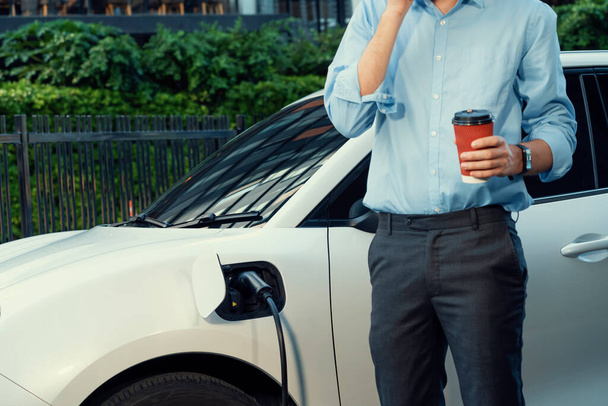 Progressive eco-friendly concept of parking EV car at public electric-powered charging station in city with blur background of businessman leaning on recharging-electric vehicle with coffee. - Φωτογραφία, εικόνα